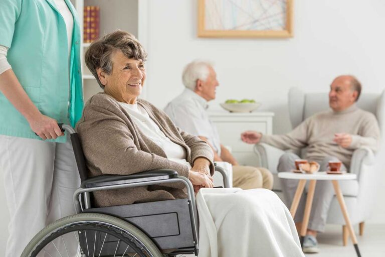 Older couple sharing a meal in an accessible assisted living apartment while a caregiver provides discreet support.